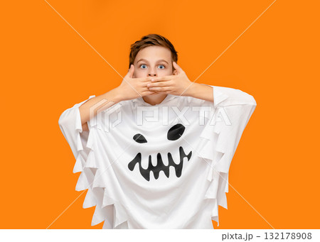 Smiling boy dressed as ghost is pointing upward, standing in front of vibrant orange background Halloween. 132178908