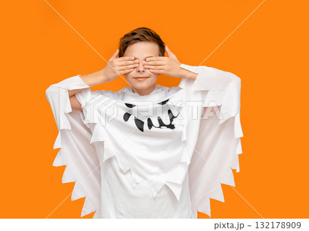 Smiling boy dressed as ghost is pointing upward, standing in front orange background, likely themed for Halloween. 132178909