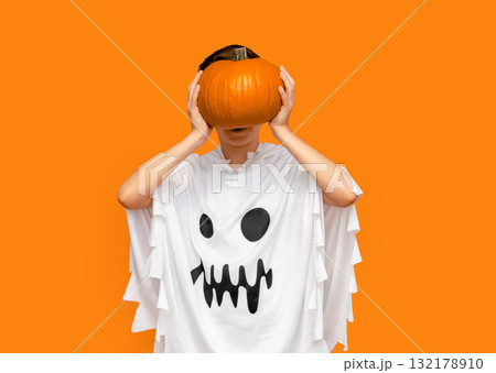 Young boy dressed in white ghost costume holds pumpkin and screams with wide-open mouth against vibrant orange background Young boy dressed in white ghost costume holds pumpkin and screams with wide-open mouth against vibrant orange background 132178910