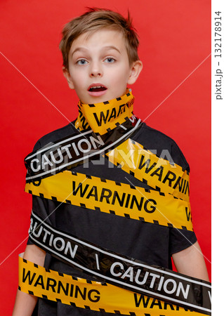 Boy stands with yellow warning tape wrapped around his body and mouth, creating dramatic visual on bold red background. 132178914