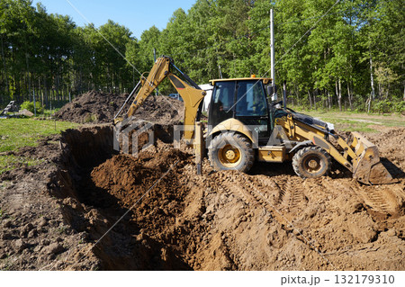 A digger operates in a rural setting, carefully digging a deep excavation for the construction of a house 132179310