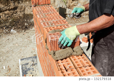 A skilled bricklayer is carefully applying mortar to red bricks, aligning them precisely to build a wall at a construction site 132179318