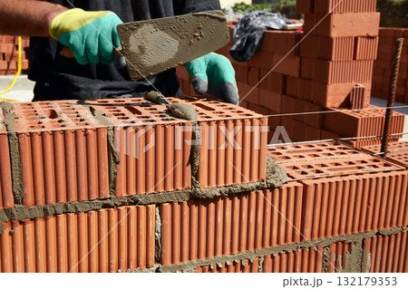 A skilled bricklayer is carefully applying mortar to red bricks, aligning them precisely to build a wall at a construction site A skilled bricklayer is carefully applying mortar to red bricks, aligning them precisely to build a wall at a construction site 132179353
