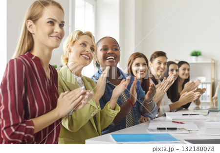 Smiling diverse group of women sitting at desk, clapping hands, supporting presentation or event. 132180078