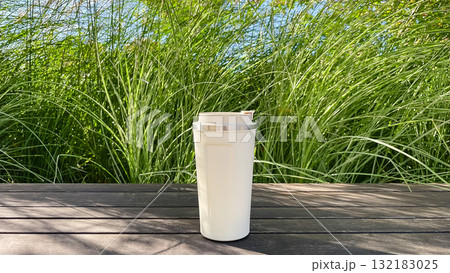 Minimalist white travel mug on wooden deck with lush green reeds. Sustainable coffee/tea in nature 132183025