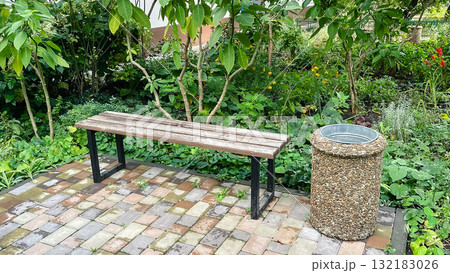Rustic park bench next to pebble trash can on patterned patio tile among colorful garden flowers 132183026