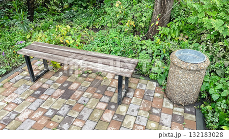 Rustic park bench next to pebble trash can on patterned patio tile among colorful garden flowers 132183028