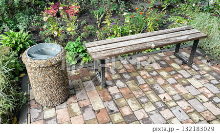 Rustic park bench next to pebble trash can on patterned patio tile among colorful garden flowers 132183029