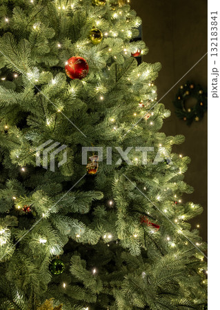 Close-up of a decorated Christmas tree with sparkling lights, red and gold ornaments, and a festive wreath blurred in the background. 132183841