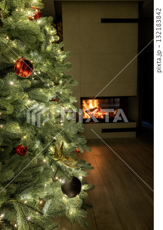 Beautiful Christmas tree with glowing lights and red ornaments standing near a cozy fireplace, creating a festive and warm holiday atmosphere. 132183842