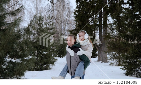 Loving couple piggyback riding through snow covered park, enjoying winter landscape and intimate playful moment together during holiday season Loving couple piggyback riding through snow covered park, enjoying winter landscape and intimate playful moment together during holiday season 132184009