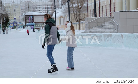 Young couple holding hands while gliding across outdoor ice rink, sharing romantic moment amid winter cityscape's festive backdrop 132184024