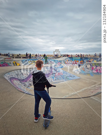 Le Havre, Normandy, France - 29 May, 2025 Rear view of a boy on a scooter looking toward the graffiti-covered skatepark bowl, at Docks Vauban Skatepark near the beach in Le Havre 132184904