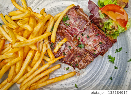 A large steak frites plate, featuring a well-seared skirt steak piled next to a generous portion of golden french fries. The meal is garnished with parsley and a simple salad of lettuce and tomato A large steak frites plate, featuring a well-seared skirt steak piled next to a generous portion of golden french fries. The meal is garnished with parsley and a simple salad of lettuce and tomato 132184915
