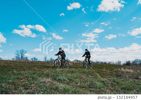 Two cyclists enjoy a sunny day biking across a grassy hill under a bright blue sky. 132184957