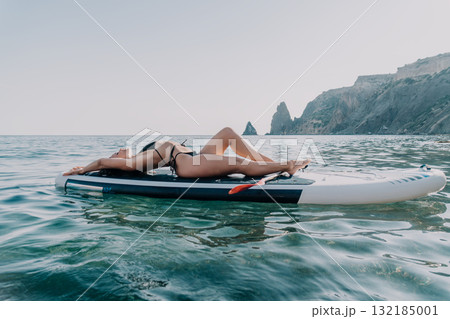 Woman Relaxing Paddleboard Sea - A woman sunbathes on a paddleboard in the ocean near cliffs. Woman Relaxing Paddleboard Sea - A woman sunbathes on a paddleboard in the ocean near cliffs. 132185001