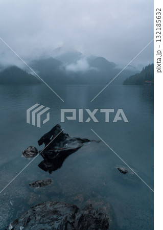 Serene lake landscape with misty mountains and rocky shoreline under cloudy sky 132185632