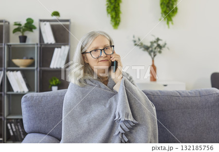 Serious senior woman wrapped in blanket sitting on sofa, talking on mobile phone and feeling unwell. Serious senior woman wrapped in blanket sitting on sofa, talking on mobile phone and feeling unwell. 132185756
