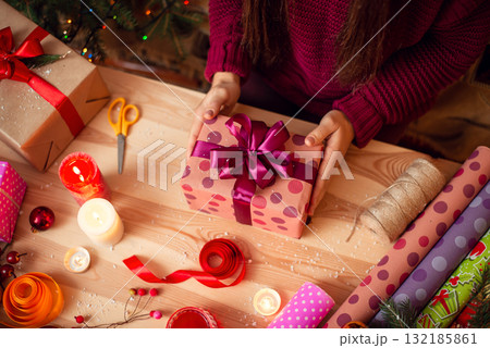 Young woman holding ready wrapped present above the table with different decorations around. Wrapping presents for close people for Christmas holidays 132185861