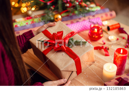 Girl checks present after wrapping it. Preparing gifts for friends and relatives in Christmas eve. Wrapping paper, candles, different decorations on the table Girl checks present after wrapping it. Preparing gifts for friends and relatives in Christmas eve. Wrapping paper, candles, different decorations on the table 132185863
