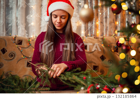 Portrait of a woman in red santa hat, sitting in sofa and making christmas wreath. Festive Christmas atmosphere, homemade decorations 132185864