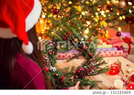 Shot of a girl holding handmade christmas wreath with bright decorated Christmas tree on the background. Christmas preparations, self made decorations 132185869