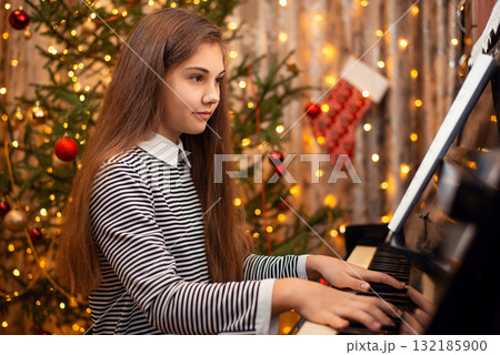 Young girl in a dress with long hair sitting near the piano and playing, Christmas tree on the background. Concept of the New Year, family holidays. 132185900