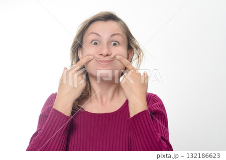 Woman forcing a smile. A woman with a maroon sweater on a white background uses her fingers to force the corners of her mouth into a smile. Woman forcing a smile. A woman with a maroon sweater on a white background uses her fingers to force the corners of her mouth into a smile. 132186623