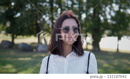 A woman with sunglasses is standing in a green outdoor space, wearing a white shirt. She looks happy as she enjoys the sunny weather surrounded by trees and rocks. A woman with sunglasses is standing in a green outdoor space, wearing a white shirt. She looks happy as she enjoys the sunny weather surrounded by trees and rocks. 132187042