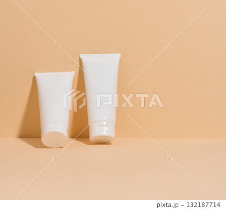 Two white cosmetic tubes without markings on a minimalist beige background. Two white cosmetic tubes without markings on a minimalist beige background. 132187714
