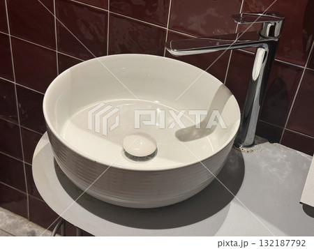 Close-up of round white sink and chrome faucet against glossy red tiled wall. Modern bathroom interior, simplicity, and geometric contrast in contemporary design. Close-up of round white sink and chrome faucet against glossy red tiled wall. Modern bathroom interior, simplicity, and geometric contrast in contemporary design. 132187792