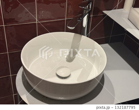 Close-up of round white sink and chrome faucet against glossy red tiled wall. Modern bathroom interior, simplicity, and geometric contrast in contemporary design. Close-up of round white sink and chrome faucet against glossy red tiled wall. Modern bathroom interior, simplicity, and geometric contrast in contemporary design. 132187795