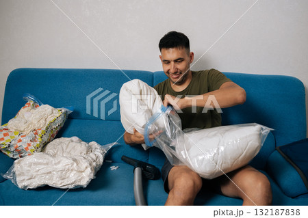Smiling young man packing white pillows and bedding into vacuum storage bags sitting on blue sofa in living room, getting ready for moving or tidying up. Concept of space saving storage. Smiling young man packing white pillows and bedding into vacuum storage bags sitting on blue sofa in living room, getting ready for moving or tidying up. Concept of space saving storage. 132187838