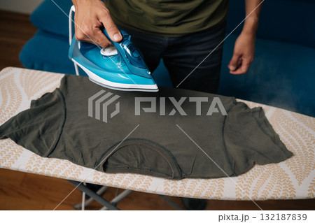 Close-up cropped shot of man carefully ironing dark green t-shirt on ironing board, using steam iron to remove wrinkles and achieve crisp at home. Concept of household chores and housework. 132187839