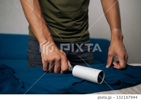 Close-up cropped shot of unrecognizable man using sticky lint roller to remove dust and lint from blue t-shirt on ironing board, preparing to iron and care for clothes at home. Close-up cropped shot of unrecognizable man using sticky lint roller to remove dust and lint from blue t-shirt on ironing board, preparing to iron and care for clothes at home. 132187944