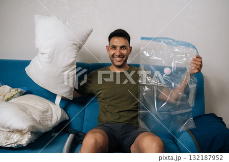 Portrait of cheerful man packing white pillows and bedding into vacuum storage bags sitting on blue sofa in living room, getting ready for moving or tidying up. Concept of space saving storage. 132187952