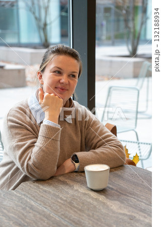 Smiling woman enjoys coffee at a cozy cafe near a large window, wearing a beige sweater and blue shirt Smiling woman enjoys coffee at a cozy cafe near a large window, wearing a beige sweater and blue shirt 132188934
