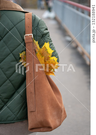 Close-up of a person wearing a green quilted jacket and brown suede bag with yellow autumn leaves tucked under the strap Close-up of a person wearing a green quilted jacket and brown suede bag with yellow autumn leaves tucked under the strap 132188941