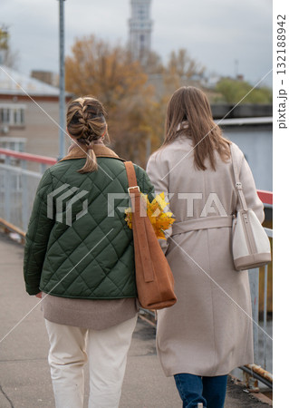 Two women walk outdoors on an overcast autumn day, one wearing a green quilted jacket and brown suede bag with yellow leaves Two women walk outdoors on an overcast autumn day, one wearing a green quilted jacket and brown suede bag with yellow leaves 132188942