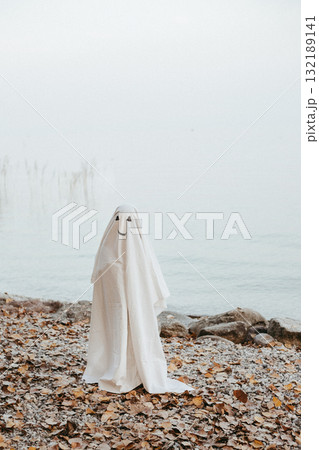 little child dressed up as a ghost standing on the beach of the lake little child dressed up as a ghost standing on the beach of the lake 132189141