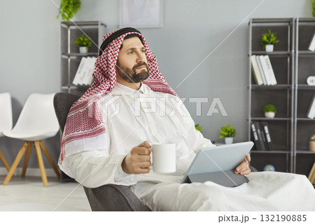 Calm executive in arab traditional dress holding coffee and tablet in casual office 132190885