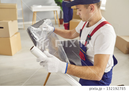 Portrait of young male mover packing furniture with plastic wrap in the living room 132192785