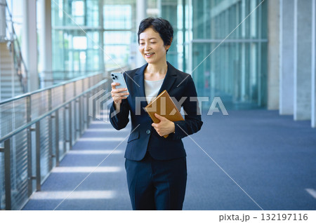スマートフォンを見ながらオフィスを歩く笑顔のビジネスウーマン (キャリア、ミドル、1人) 132197116