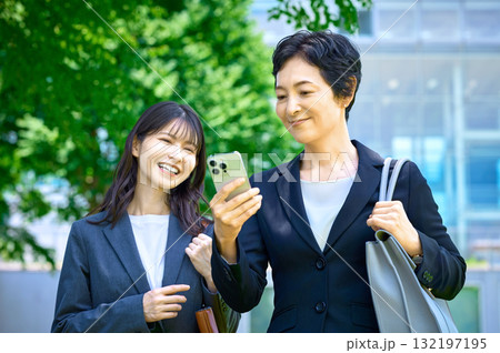 屋外でスマートフォンを見ながら笑顔で歩く二人のビジネスウーマン 132197195