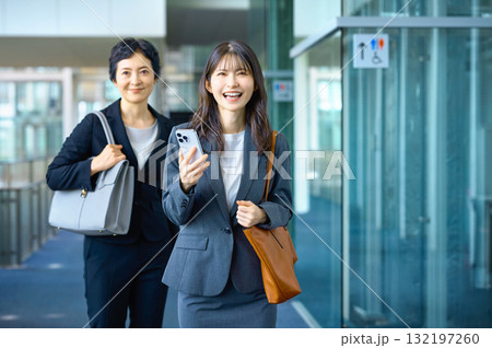 スマートフォンを持って笑顔でオフィスを歩く二人のビジネスウーマン (若手とミドル、キャリア、2人) 132197260