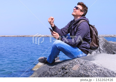 Man traveler is happy about getting great news on smartphone sits at sea beach. 132202416