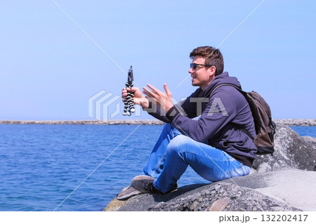 Travel blogger man shoots video on smartphone sits on stone at sea beach. Travel blogger man shoots video on smartphone sits on stone at sea beach. 132202417
