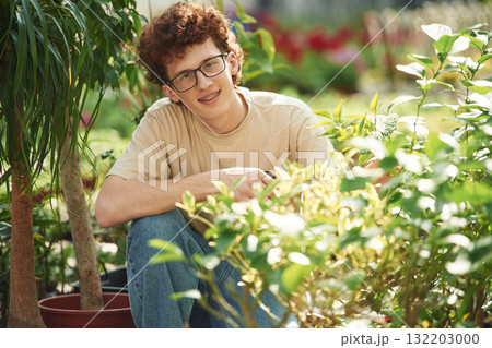Growing flowers. Conception of watering. Young man with curly hair and in glasses is in greenhouse 132203000