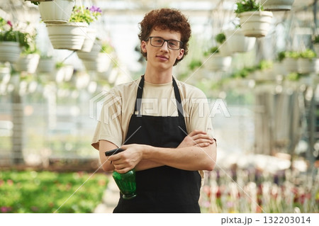 Arms crossed. With watering bottle. Young man with curly hair and in glasses is in greenhouse Arms crossed. With watering bottle. Young man with curly hair and in glasses is in greenhouse 132203014