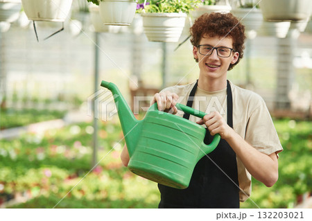 Green watering can in hands. Young man with curly hair and in glasses is in greenhouse 132203021